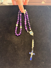 A hand holds Redemption Jewelry’s LR004 Long Amethyst & Howlite Rosary, featuring purple and white beads, a blue and silver medallion, and a cross with blue inlay, set against a black background.