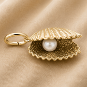 The Shell With Pearl Charm- Gold Plated by Rembrandt Charms, featuring a gold-plated seashell design with a lustrous white pearl inside, is displayed on a beige fabric background, capturing the elegance of ocean-inspired jewelry.