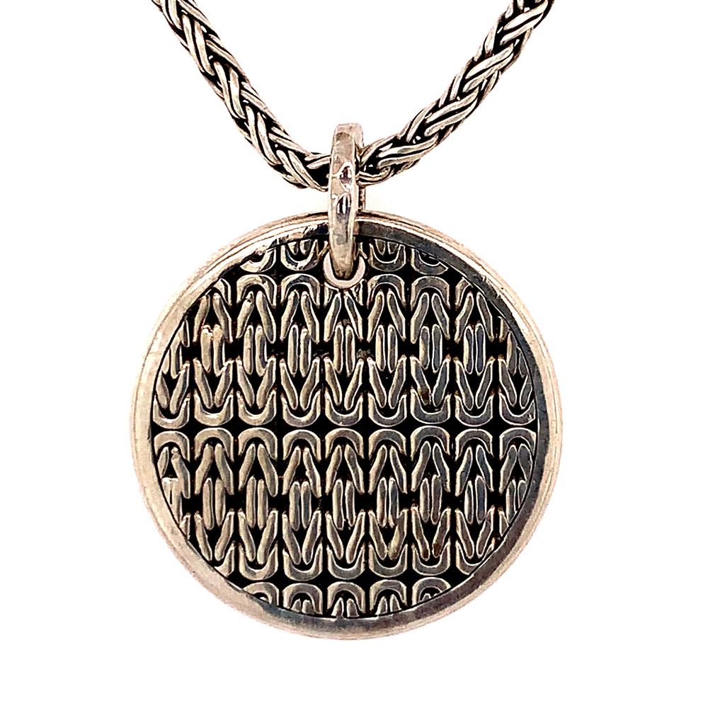 The Lois Hill Sterling Silver Gents Reversible Necklace boasts an intricate, woven metallic design on its circular pendant, which elegantly hangs from a twisted sterling silver chain. The pendant features a detailed pattern reminiscent of interlocking links or scales, providing a textured and luxurious appearance that reflects the expert craftsmanship throughout.