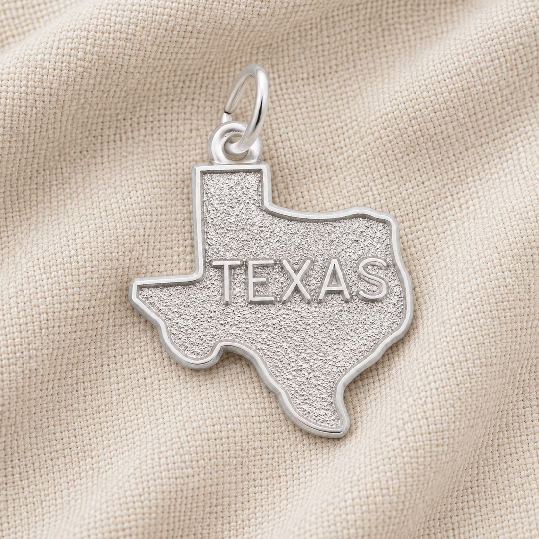 A Rembrandt Charms Texas State Map Charm, crafted in sterling silver and engraved with “TEXAS,” rests on a textured beige fabric background.
