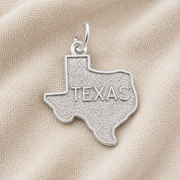 A Rembrandt Charms Texas State Map Charm, crafted in sterling silver and engraved with “TEXAS,” rests on a textured beige fabric background.