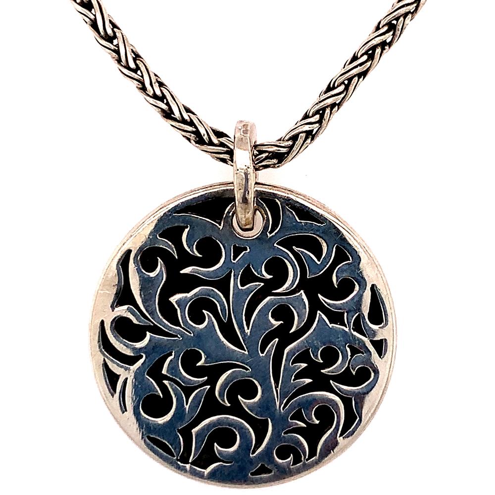 Presenting the Sterling Silver Gents Reversible Necklace by Lois Hill: a sterling silver necklace featuring an intricately cut-out floral design on a circular pendant. Hanging gracefully from a woven chain, the design stands out against a dark background. This versatile piece doubles as a reversible necklace, making it perfect for gents seeking both style and versatility.