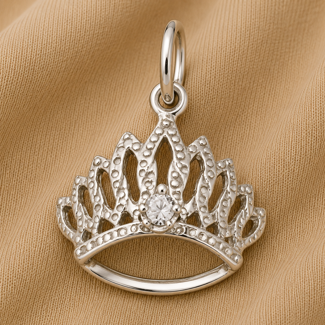 The Tiara With Birthstone Charm (April) by Rembrandt Charms, crafted in sterling silver with intricate crown-shaped detailing and a sparkling central gem, reminiscent of a regal jewelry piece or April birthstone tiara, rests on textured beige fabric.