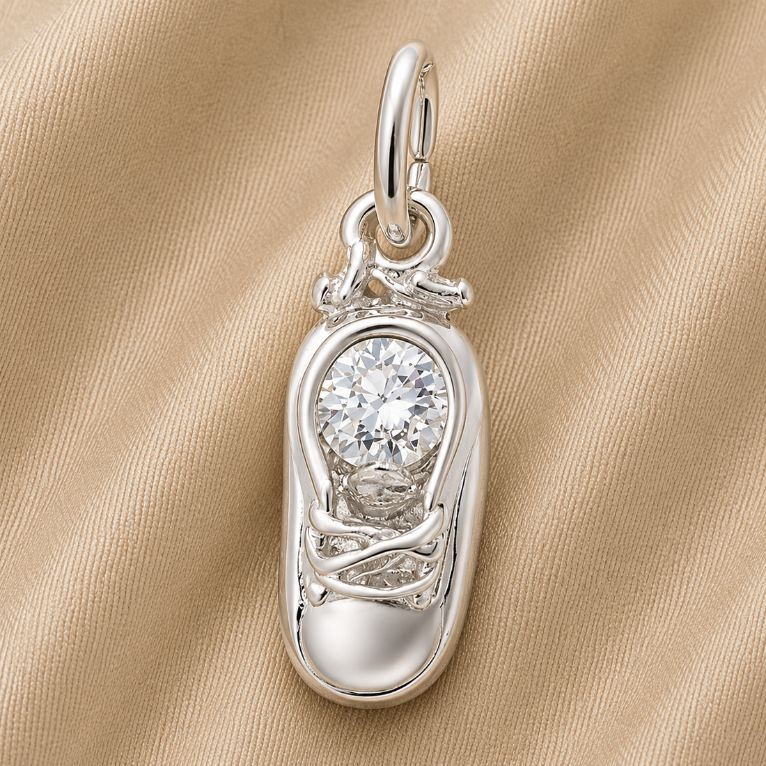 The Rembrandt Charms Baby Shoe Birthstone (April) in sterling silver features a sneaker-shaped pendant accented with a sparkling round gemstone, creating a charming April birthstone keepsake, beautifully displayed on a beige textured fabric background.