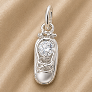 The Rembrandt Charms Baby Shoe Birthstone (April) in sterling silver features a sneaker-shaped pendant accented with a sparkling round gemstone, creating a charming April birthstone keepsake, beautifully displayed on a beige textured fabric background.