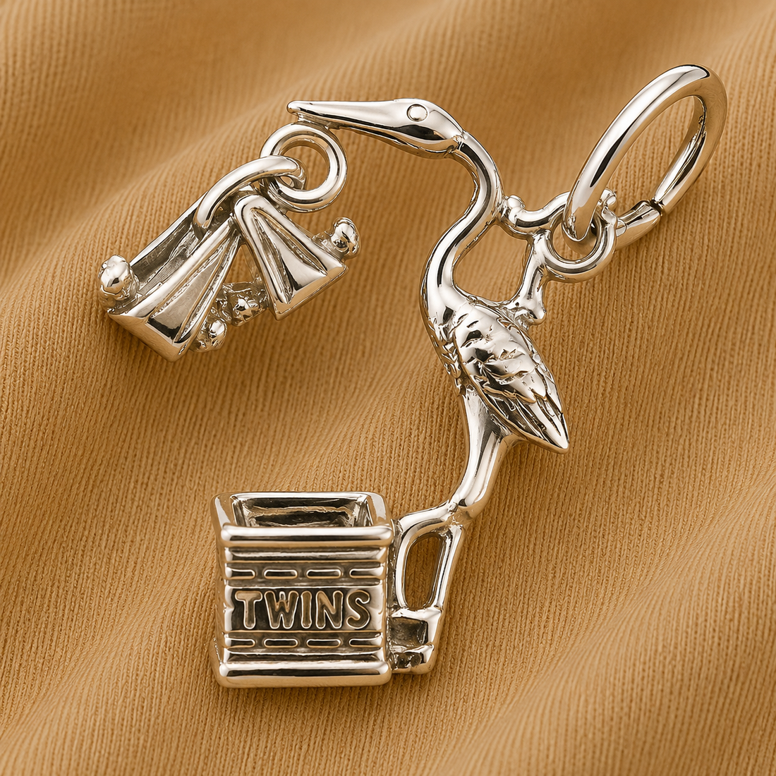 The Rembrandt Charms Twins Stork Charm in sterling silver features a stork holding a bundle in its beak, standing beside a basket engraved with “TWINS,” all displayed on a tan fabric background—an endearing piece of stork jewelry that makes a meaningful gift for new parents or a sentimental twins charm.