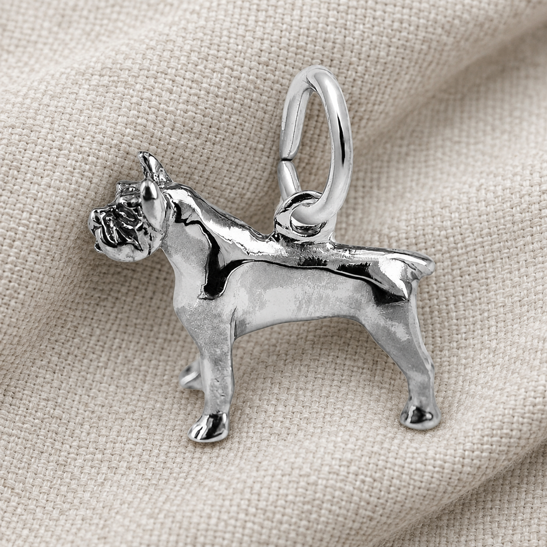 A gleaming Pitbull Dog Charm in sterling silver from Rembrandt Charms rests on a textured beige fabric background—an ideal keepsake for dog lovers.