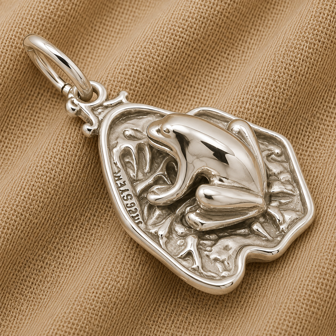 A gleaming sterling silver charm from Rembrandt Charms, the “Frog on Lily Pad Charm,” featuring intricate leaf details reminiscent of a delicate frog resting among lily pads, displayed atop textured beige fabric.