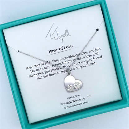 The Paws of Love Sterling Silver Charm Necklace by T Jazelle features a heart-shaped pendant engraved with paw prints, comes in a turquoise box, and includes a display card reading: “Paws of Love,” symbolizing cherished memories.