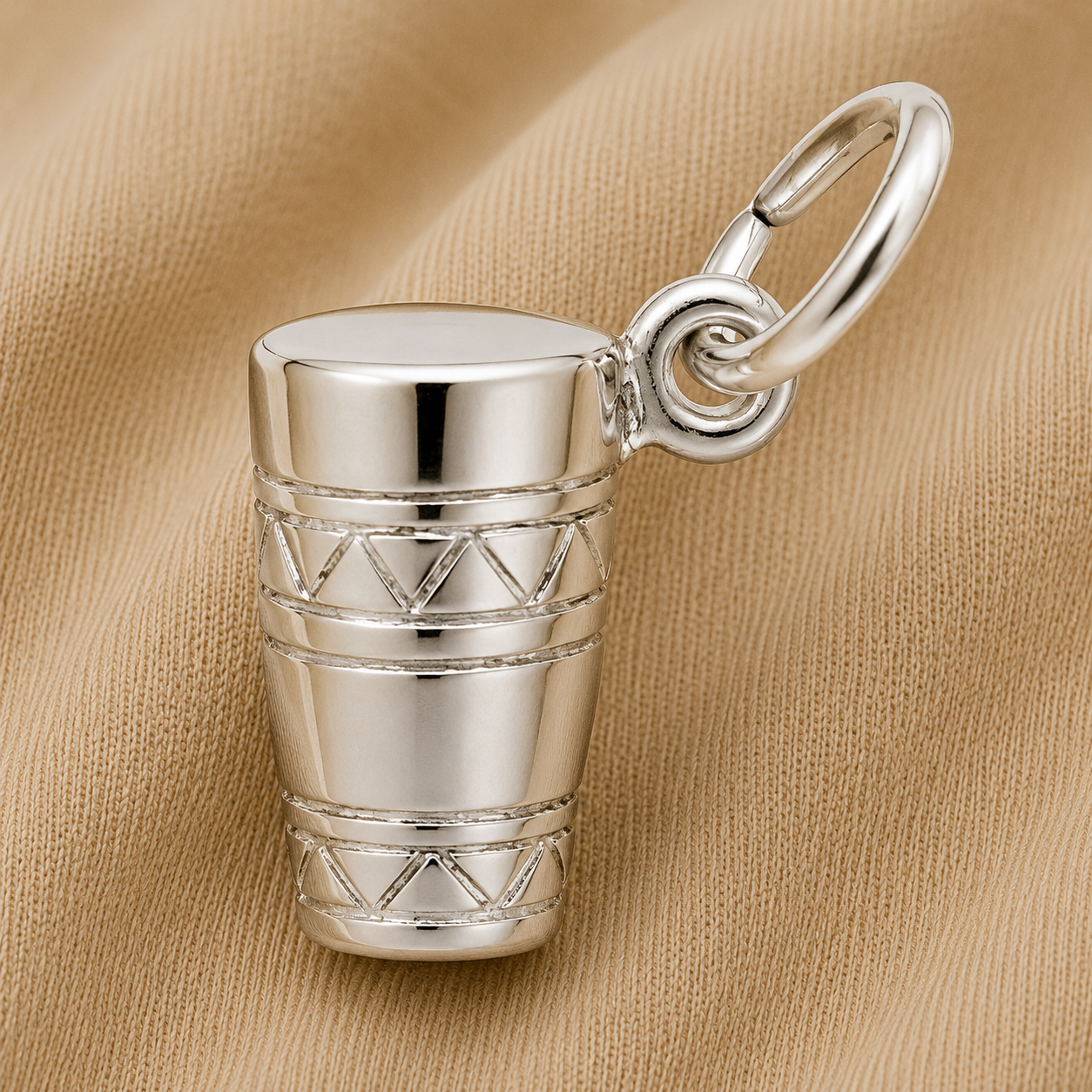 The Rembrandt Charms Bongo Drum Charm in sterling silver, adorned with engraved geometric patterns, rests on a beige, textured fabric background—an elegant piece of music-themed jewelry.