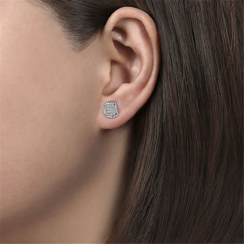 Close-up of a person's ear wearing Gabriel & Co's 925 Sterling Silver White Sapphire and Rope Frame Stud Earrings *Ham. These square-shaped studs, featuring a sparkling design that appears to be encrusted with small white sapphires, add an elegant touch to any look. The person has straight, dark brown hair.