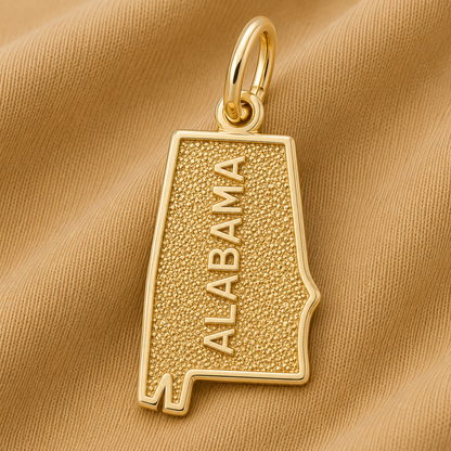 A gold-plated Alabama Charm by Rembrandt Charms, shaped like the state of Alabama with “ALABAMA” engraved vertically and displayed on a beige textured fabric background—this elegant piece makes the perfect Alabama jewelry for showcasing your state pride.