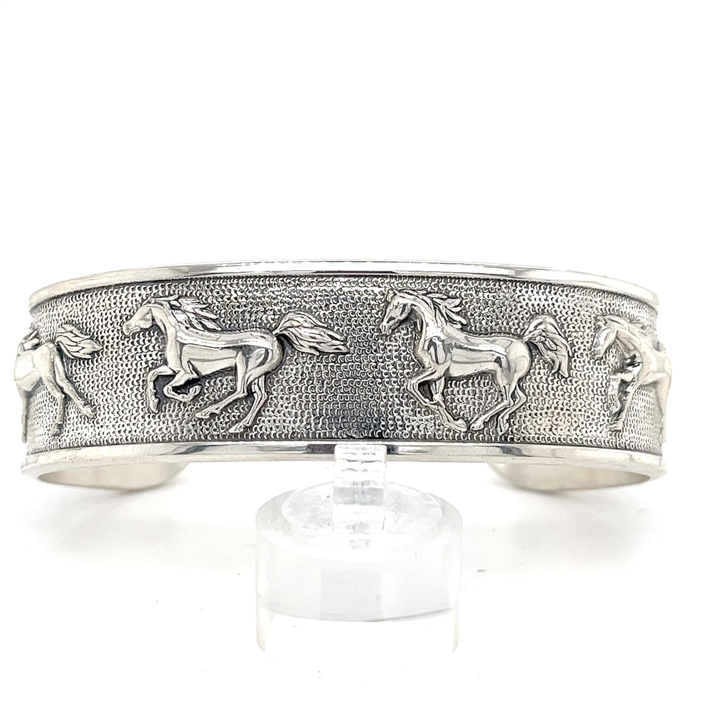 The Kabana SS Large Horse Bracelet Cuff features a raised galloping horse design on textured silver, elegantly displayed on a clear stand with a white backdrop.