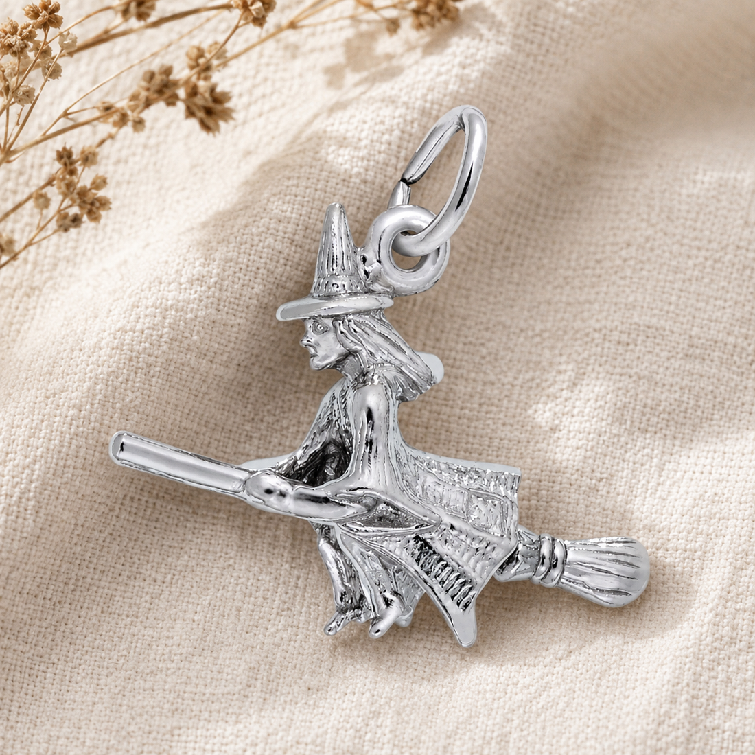 The Witch Charm- Sterling Silver by Rembrandt Charms is displayed on a beige fabric background with dried flowers in the corner—an enchanting piece of Halloween jewelry perfect for adding a touch of magic to your collection.