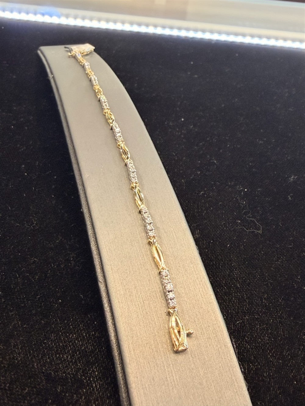 The Station Diamond and Gold Tennis Bracelet by Ryan Gems, crafted in 18k white and yellow gold, is showcased on a curved gray stand against a black background, with bright lighting accentuating its sparkling diamonds.