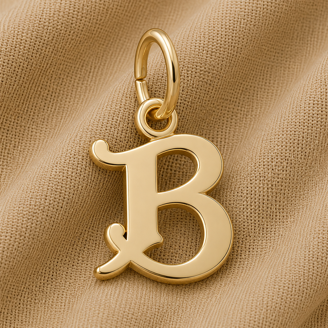 A gold-plated Curly Initial Charm (B) by Rembrandt Charms rests on beige textured fabric, featuring a top loop for easy attachment to a chain or bracelet. This elegant initial charm is perfect as keepsake jewelry or as a personalized accent, adding both meaning and style to any collection.