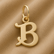A gold-plated Curly Initial Charm (B) by Rembrandt Charms rests on beige textured fabric, featuring a top loop for easy attachment to a chain or bracelet. This elegant initial charm is perfect as keepsake jewelry or as a personalized accent, adding both meaning and style to any collection.