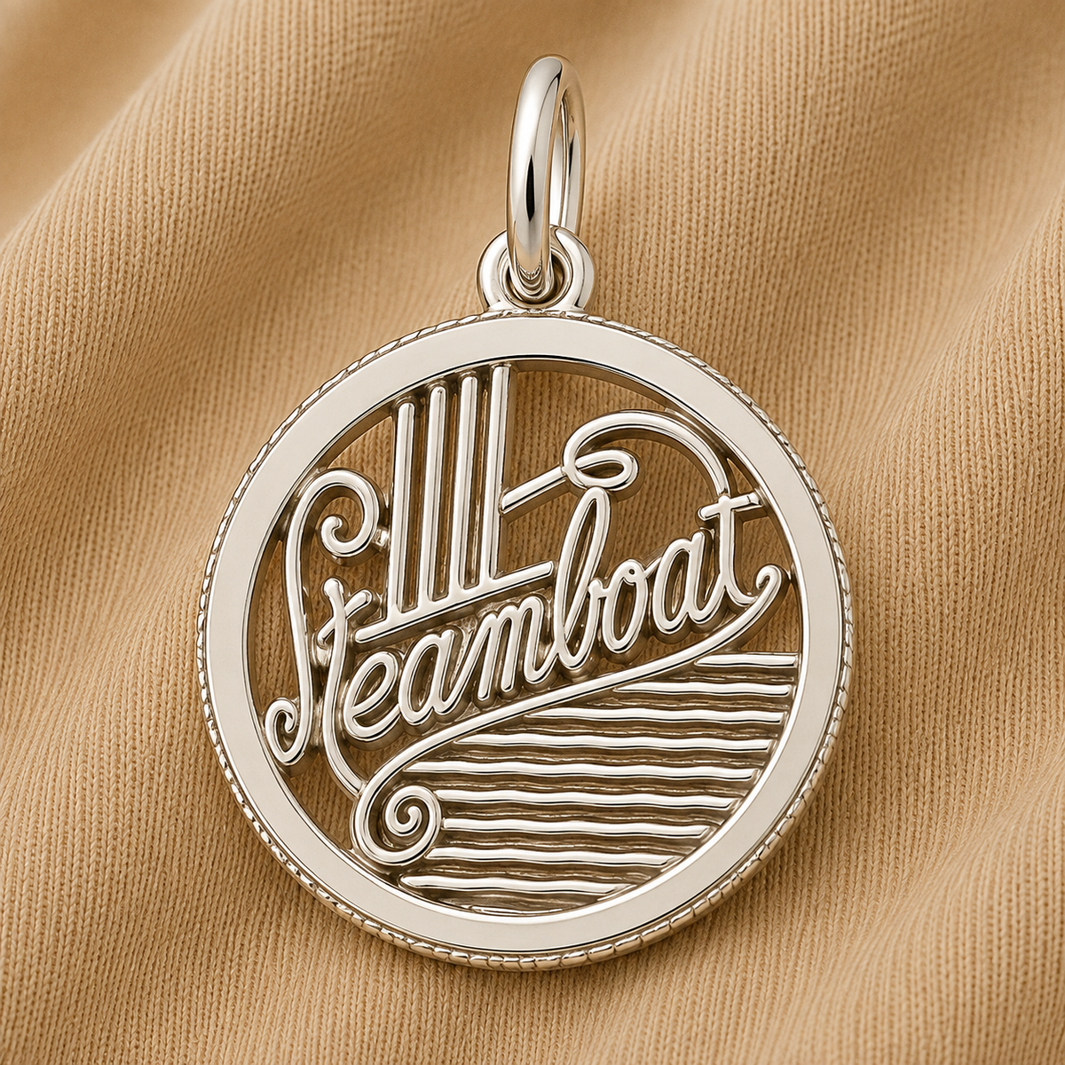 A Rembrandt Charms Steamboat Charm in sterling silver rests on tan fabric, featuring the word “Steamboat” in elegant script with stylized steam and waves inside the round pendant—a perfect piece of travel jewelry for those who cherish mountain adventure keepsakes.