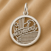 A Rembrandt Charms Steamboat Charm in sterling silver rests on tan fabric, featuring the word “Steamboat” in elegant script with stylized steam and waves inside the round pendant—a perfect piece of travel jewelry for those who cherish mountain adventure keepsakes.