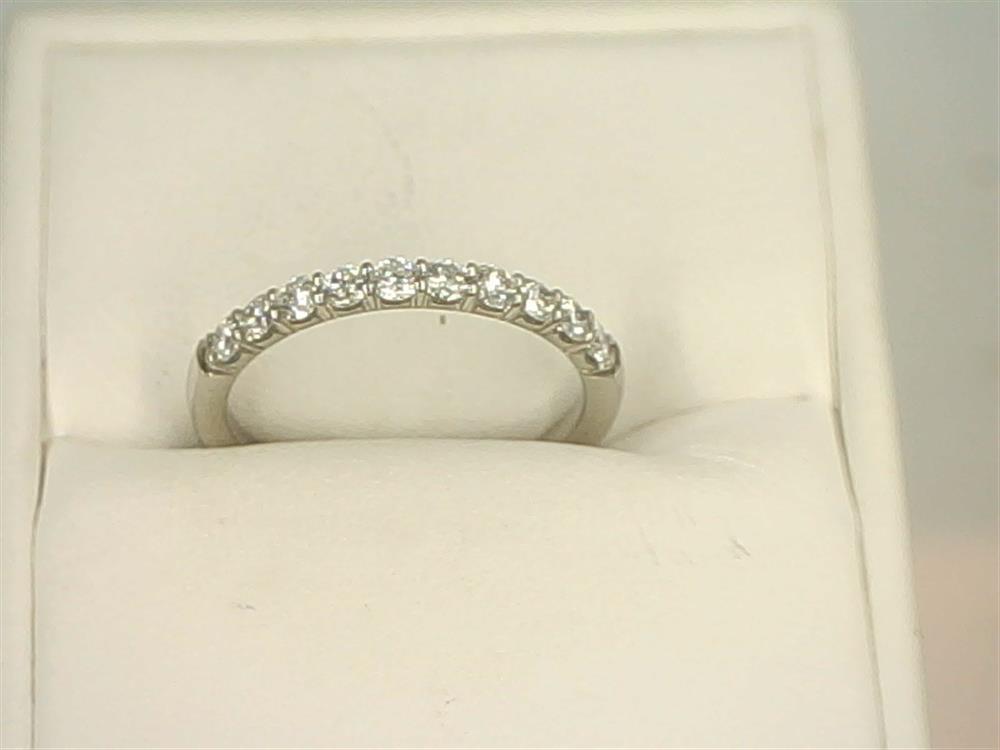 A 14K White Gold .4000ctw Round Cut Diamond Anniversary Band by Benchmark Rings is displayed on a white cushioned stand. The ring, adorned with a row of small, sparkling lab-grown diamonds set in a straight line, is showcased in a close-up, highlighting the intricate detailing and brilliance of the stones.