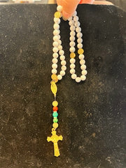 A hand holds the LR001 Long Howlite Rosary by Redemption Jewelry, featuring white beads, gold accents, a gold crucifix, and colored beads near the bottom, all set against a dark backdrop.