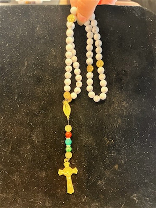 A hand holds the LR001 Long Howlite Rosary by Redemption Jewelry, featuring white beads, gold accents, a gold crucifix, and colored beads near the bottom, all set against a dark backdrop.