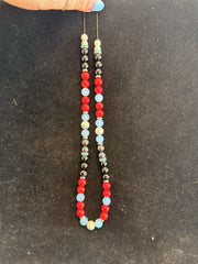 A hand with blue nail polish holds Redemption Jewelry’s NK001 necklace, featuring Howlite, Obsidian, Red Jasper, Blue Chalcedony, and Hematite beads in a colorful pattern against a dark surface.
