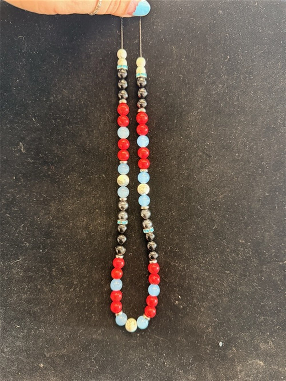 A hand with blue nail polish holds Redemption Jewelry’s NK001 necklace, featuring Howlite, Obsidian, Red Jasper, Blue Chalcedony, and Hematite beads in a colorful pattern against a dark surface.