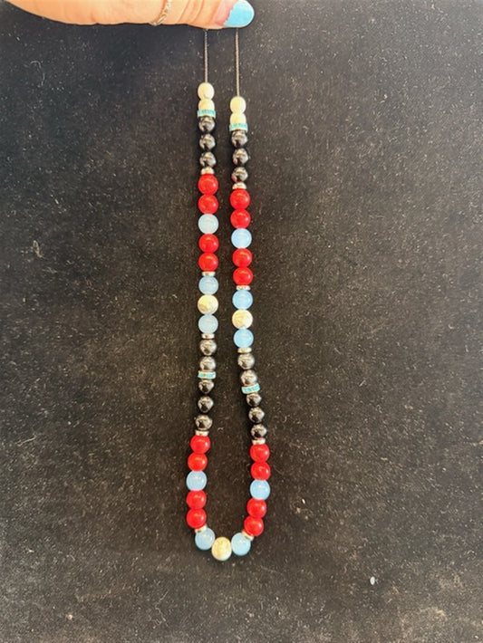 A hand with blue nail polish holds Redemption Jewelry’s NK001 necklace, featuring Howlite, Obsidian, Red Jasper, Blue Chalcedony, and Hematite beads in a colorful pattern against a dark surface.
