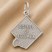 A sterling silver District of Columbia Charm by Rembrandt Charms, shaped like the city and engraved with “DISTRICT OF COLUMBIA,” is displayed on a textured beige fabric background. This distinctive piece is an ideal Washington DC keepsake for anyone wishing to carry a memory of the nation’s capital.