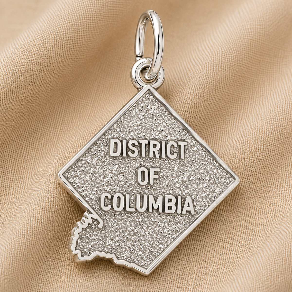 A sterling silver District of Columbia Charm by Rembrandt Charms, shaped like the city and engraved with “DISTRICT OF COLUMBIA,” is displayed on a textured beige fabric background. This distinctive piece is an ideal Washington DC keepsake for anyone wishing to carry a memory of the nation’s capital.