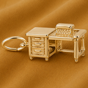 A gold-plated Typewriter and Desk Charm by Rembrandt Charms, designed with an office desk, drawers, and chair, is displayed on textured brown fabric—this distinctive accessory makes a thoughtful gift for writers or anyone who appreciates unique literary jewelry.