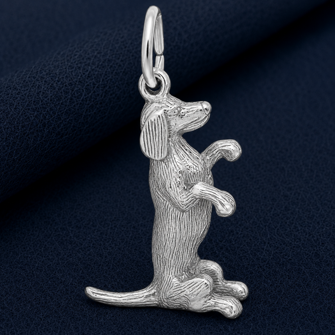The Sitting Dachshund Charm by Rembrandt Charms is crafted in sterling silver and features a dog sitting upright with raised front paws, detailed fur texture capturing every feature. Perfect as a pet keepsake, this charm is displayed on a dark blue textured surface.