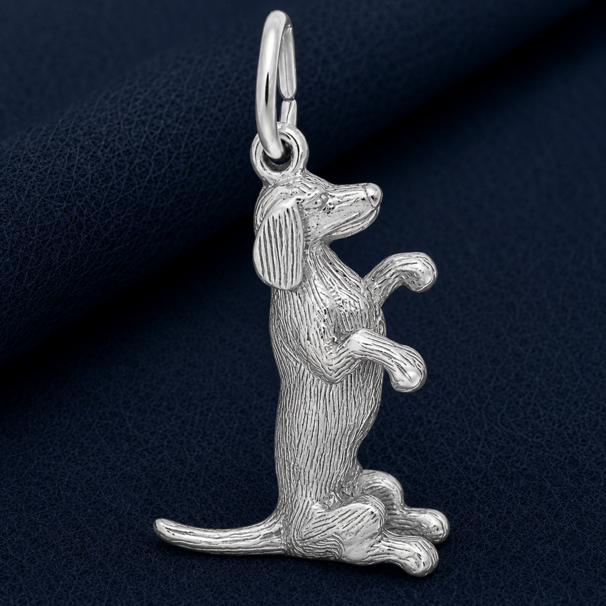 The Sitting Dachshund Charm by Rembrandt Charms is crafted in sterling silver and features a dog sitting upright with raised front paws, detailed fur texture capturing every feature. Perfect as a pet keepsake, this charm is displayed on a dark blue textured surface.