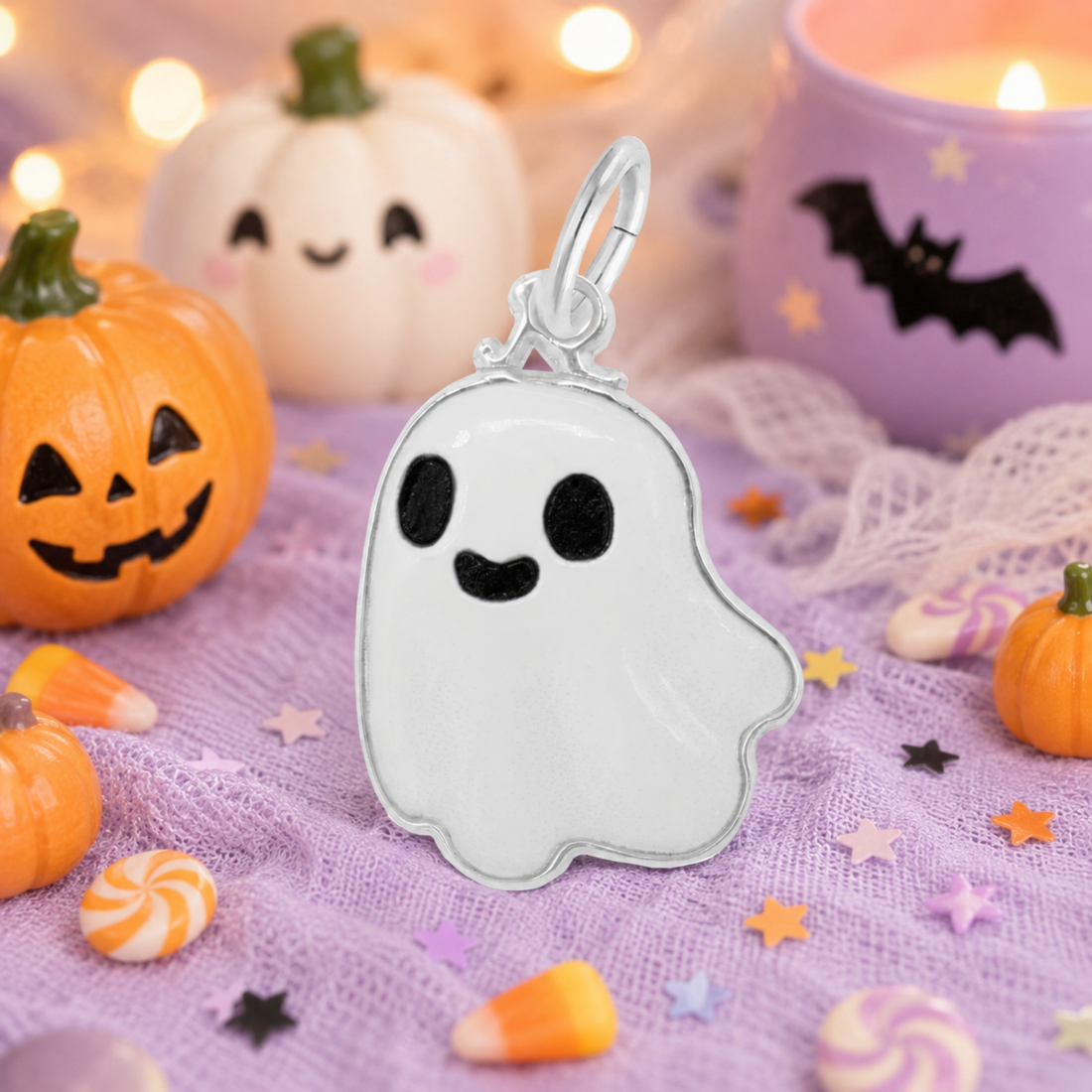 Add playful spirit to your Halloween jewelry collection with the Rembrandt Charms Painted Cute Ghost Charm in sterling silver, showcased on a festive purple cloth surrounded by pumpkins, candies, a purple candle adorned with a bat, and other cheerful Halloween decorations.