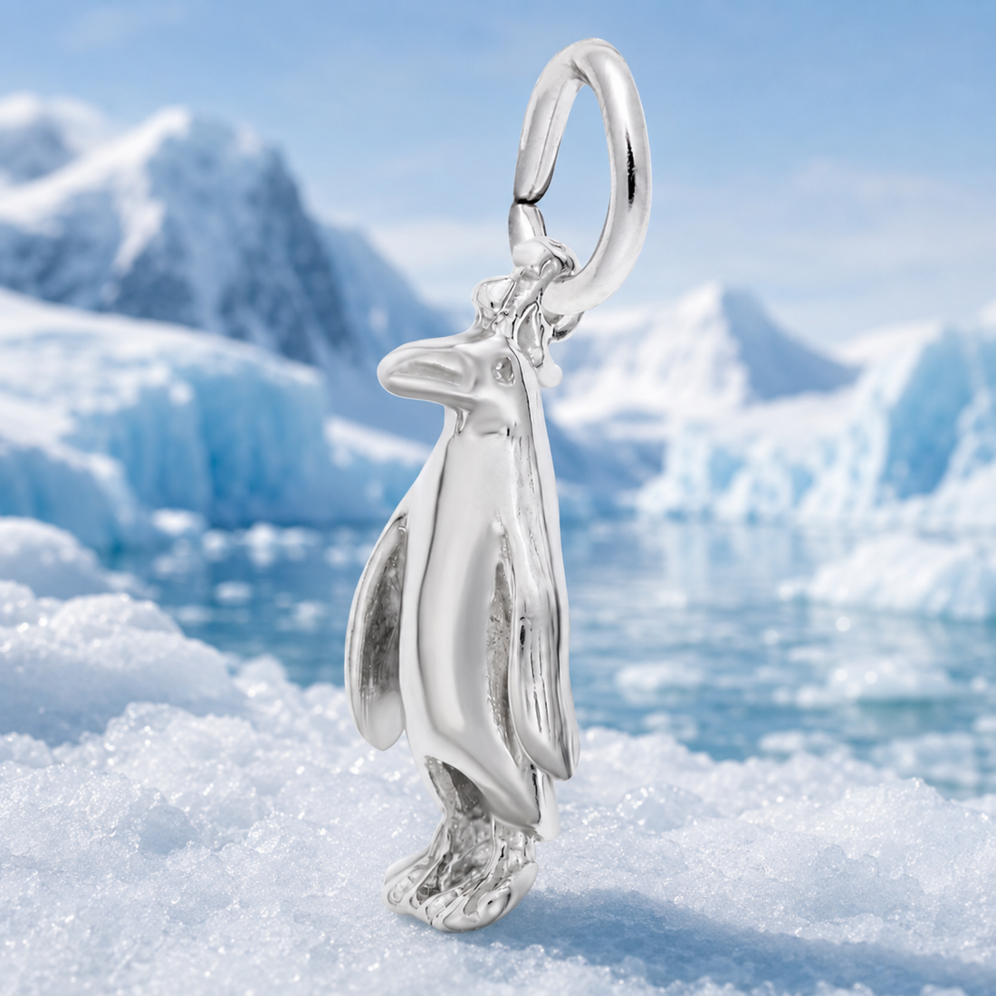 A gleaming three-dimensional Adélie Penguin – Gold Plated Charm by Rembrandt Charms stands atop icy snow, with blurred blue icebergs and snowy mountains in the background.
