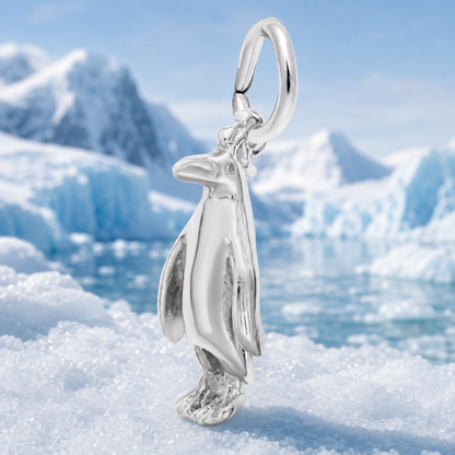 A gleaming three-dimensional Adélie Penguin – Gold Plated Charm by Rembrandt Charms stands atop icy snow, with blurred blue icebergs and snowy mountains in the background.