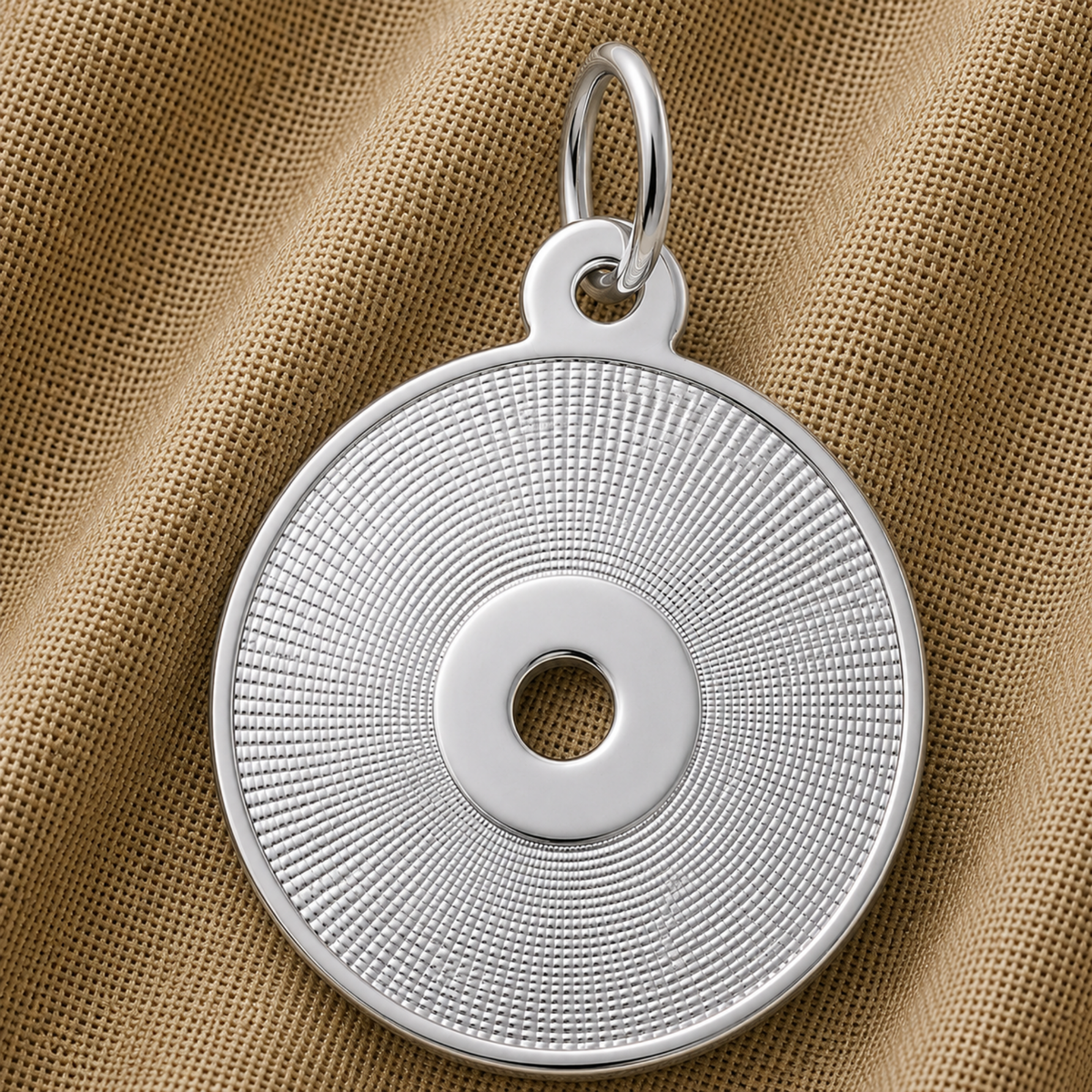 A round, sterling silver CD Charm by Rembrandt Charms—featuring a textured, radiating pattern and central hole reminiscent of a compact disc—rests elegantly on a brown textured fabric. This necklace is the perfect gift for music lovers.