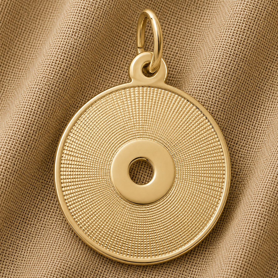The Rembrandt Charms CD Charm in sterling silver features a round, concentric textured pattern with a central hole reminiscent of a compact disc. Shown resting on a beige, textured fabric background, this elegant piece is an ideal gift for any music lover.