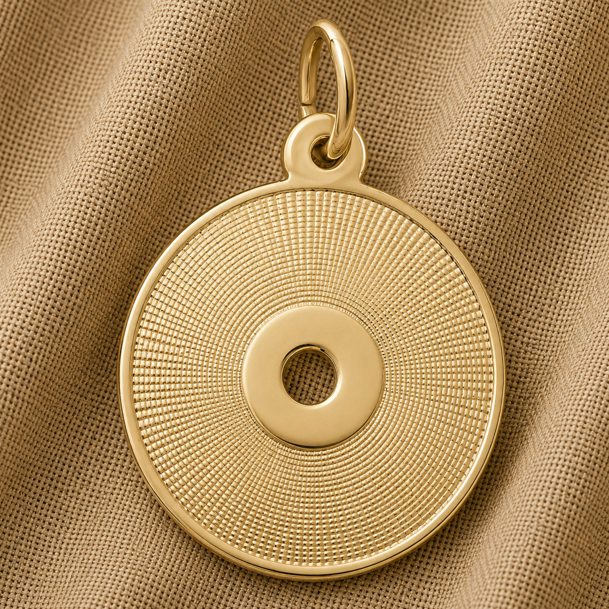 The Rembrandt Charms CD Charm in sterling silver features a round, concentric textured pattern with a central hole reminiscent of a compact disc. Shown resting on a beige, textured fabric background, this elegant piece is an ideal gift for any music lover.