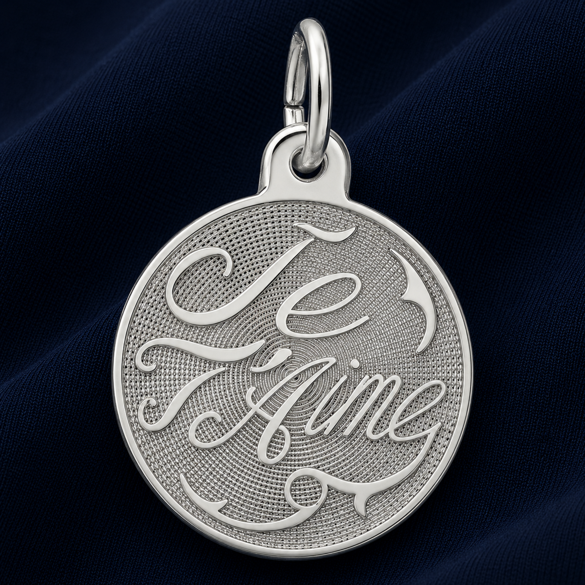 A round sterling silver Je T’aime Disc Charm by Rembrandt Charms, beautifully engraved in cursive script, displayed on a dark blue textured fabric background—a perfect French love message charm and romantic gift for someone special.