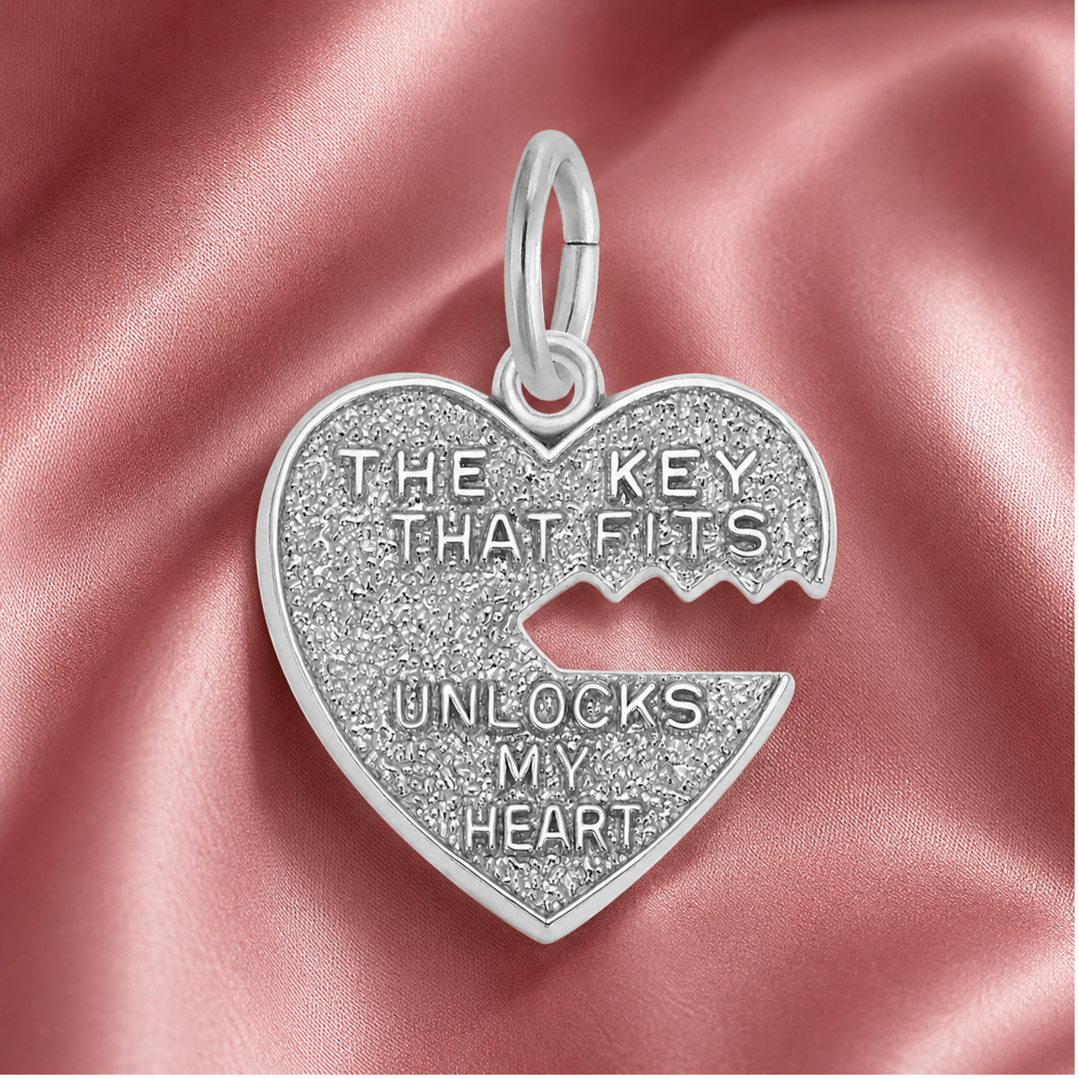 The Heart and Key charm by Rembrandt Charms, crafted in sterling silver and featuring a heart-shaped pendant with a jagged keyhole and the engraved message “THE KEY THAT FITS UNLOCKS MY HEART,” is elegantly displayed on pink satin fabric—making it the perfect piece of romantic jewelry and an unforgettable anniversary gift.