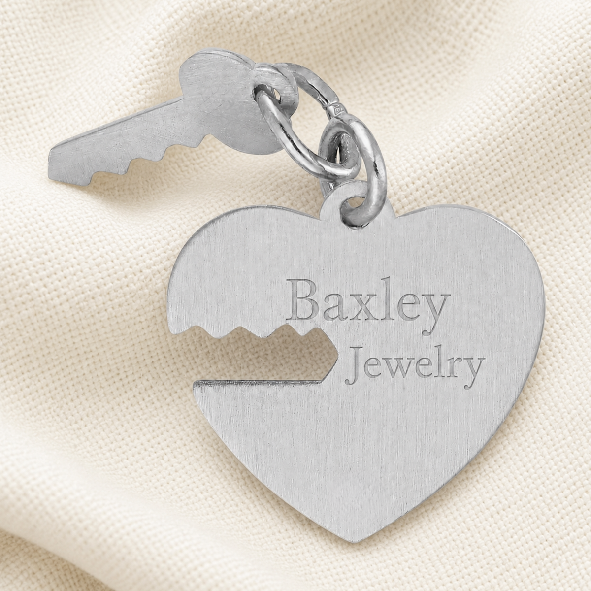 A sterling silver Heart and Key charm from Rembrandt Charms, featuring elegant jagged cutouts, rests on a cream fabric. This romantic piece makes a perfect anniversary gift.