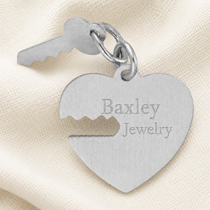 A sterling silver Heart and Key charm from Rembrandt Charms, featuring elegant jagged cutouts, rests on a cream fabric. This romantic piece makes a perfect anniversary gift.