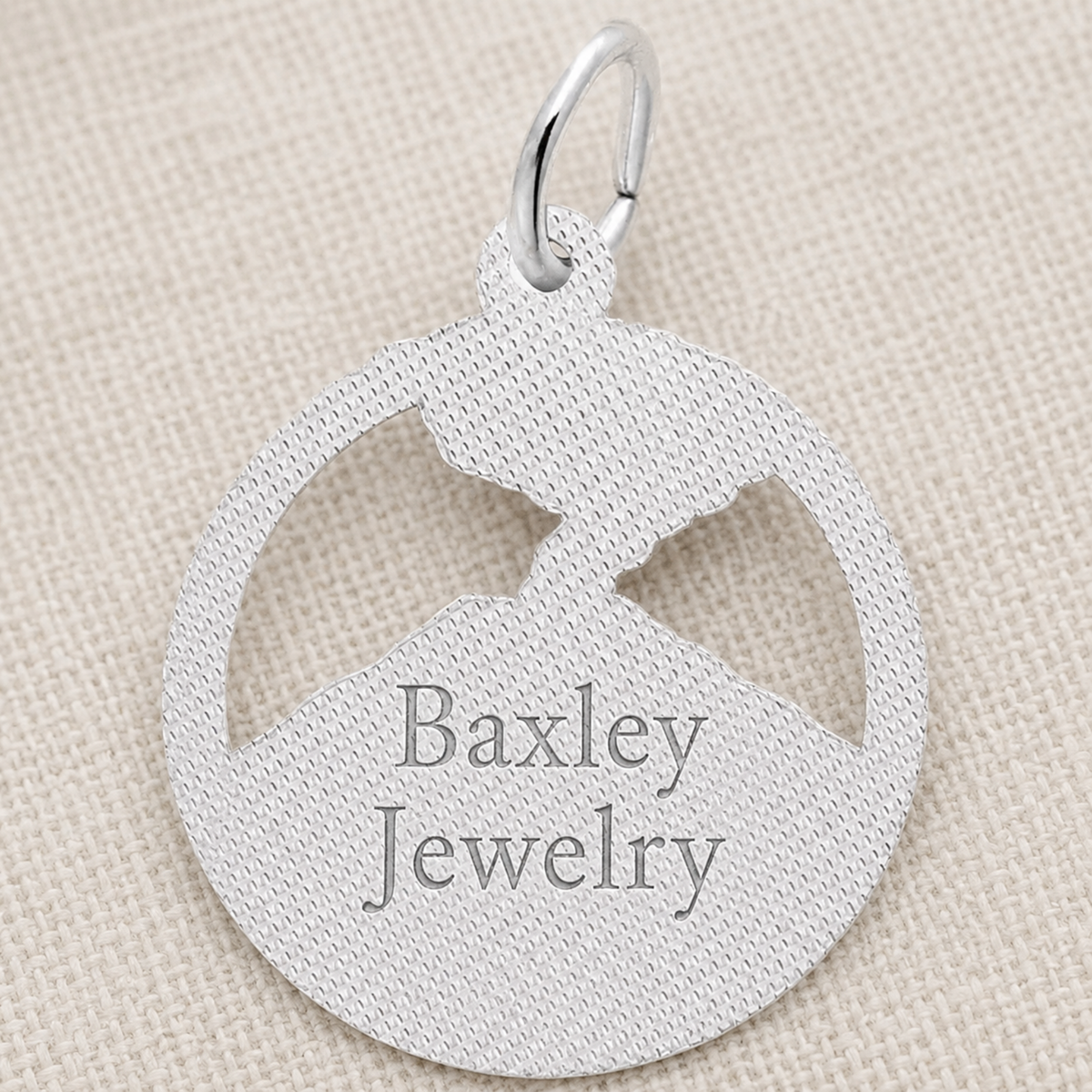 Sterling silver Mount St. Helens Disc Charm by Rembrandt Charms, featuring a round pendant with a mountain cutout, displayed on textured beige fabric. This distinctive piece is an ideal Pacific Northwest keepsake for nature enthusiasts.