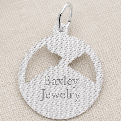 Sterling silver Mount St. Helens Disc Charm by Rembrandt Charms, featuring a round pendant with a mountain cutout, displayed on textured beige fabric. This distinctive piece is an ideal Pacific Northwest keepsake for nature enthusiasts.