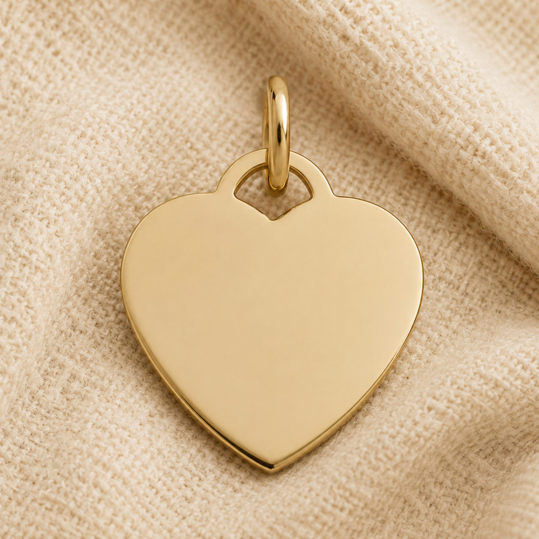 A Small Heart Classic Charm in sterling silver by Rembrandt Charms, featuring a smooth surface and perfect for engraving, rests on beige textured fabric.