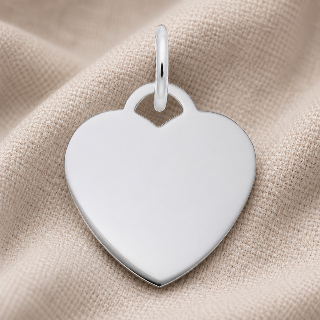 The Rembrandt Charms Small Heart Classic Charm in sterling silver features a lustrous, heart-shaped design with a loop, elegantly displayed on a soft beige fabric background—a versatile piece perfect as a heart charm or an engravable jewelry keepsake.