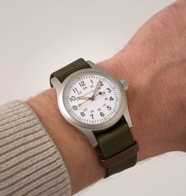 A close-up image of a person's wrist showcases the Hamilton Khaki Field Mechanical Watch with NATO Strap, featuring a white dial and green military-style fabric strap. The watch displays black Arabic numerals in a 24-hour format, has a silver casing, and evokes military heritage. The person is wearing a light beige or gray sweater.