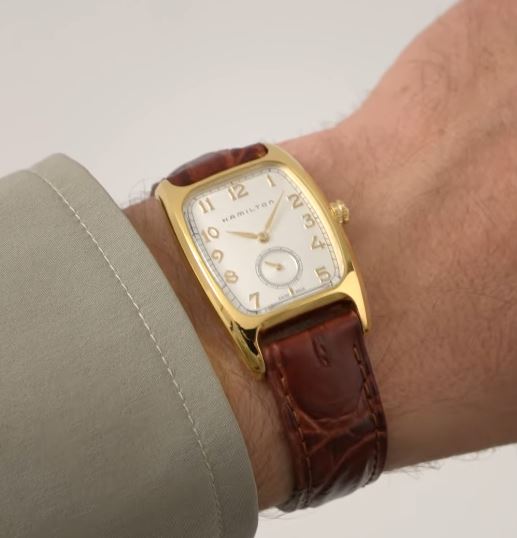 A wrist adorned with a Boulton Quartz Watch by Hamilton, showcasing its gold casing and white dial. This American classic features Arabic numerals, a brown leather strap, and is elegantly paired with a beige shirt sleeve. The "Hamilton" brand name is prominently displayed on the dial.